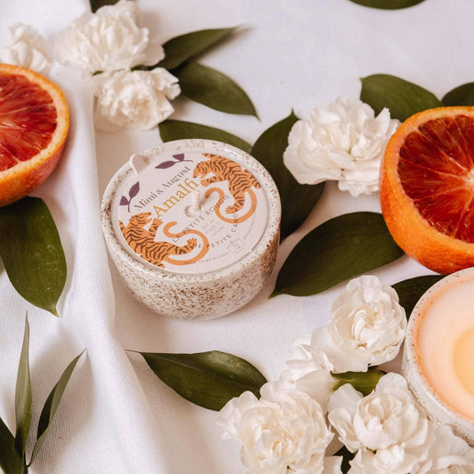 A scented candle in a speckled ceramic jar with a tiger illustration and the brand name 'TIGRE SCENTS' is displayed on a white fabric backdrop, styled with white flowers, green leaves, and sliced blood oranges.