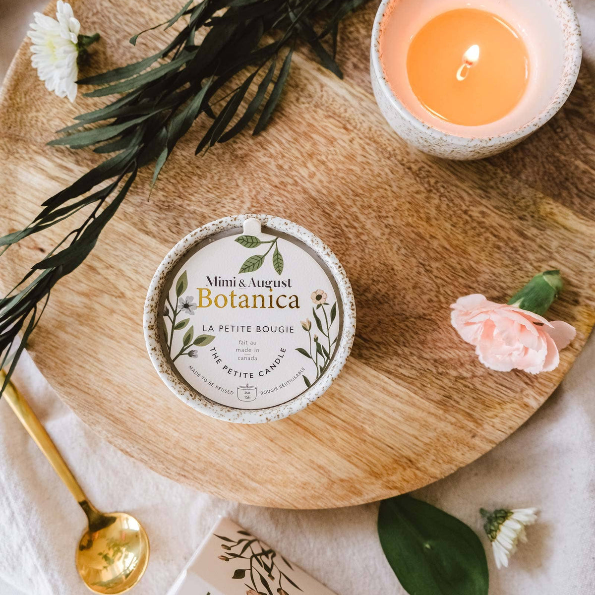 A round wooden tray holds a candle tin labeled Mimosa Baked Botanica, alongside a lit candle in a speckled bowl, greenery, flowers, a gold spoon, and a book on a soft white fabric background.