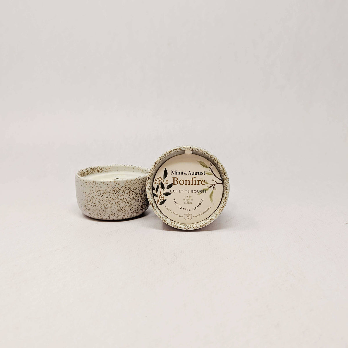 A small speckled ceramic candle jar with its lid placed beside it on a plain light background. The lid has printed text reading 'Mini & Auguste Bonfire' and 'Petite Bougie'.