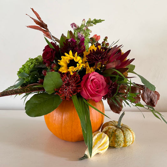 A decorative floral arrangement featuring a pumpkin and a selection of fall-colored flowers including sunflowers, roses, and various greenery.