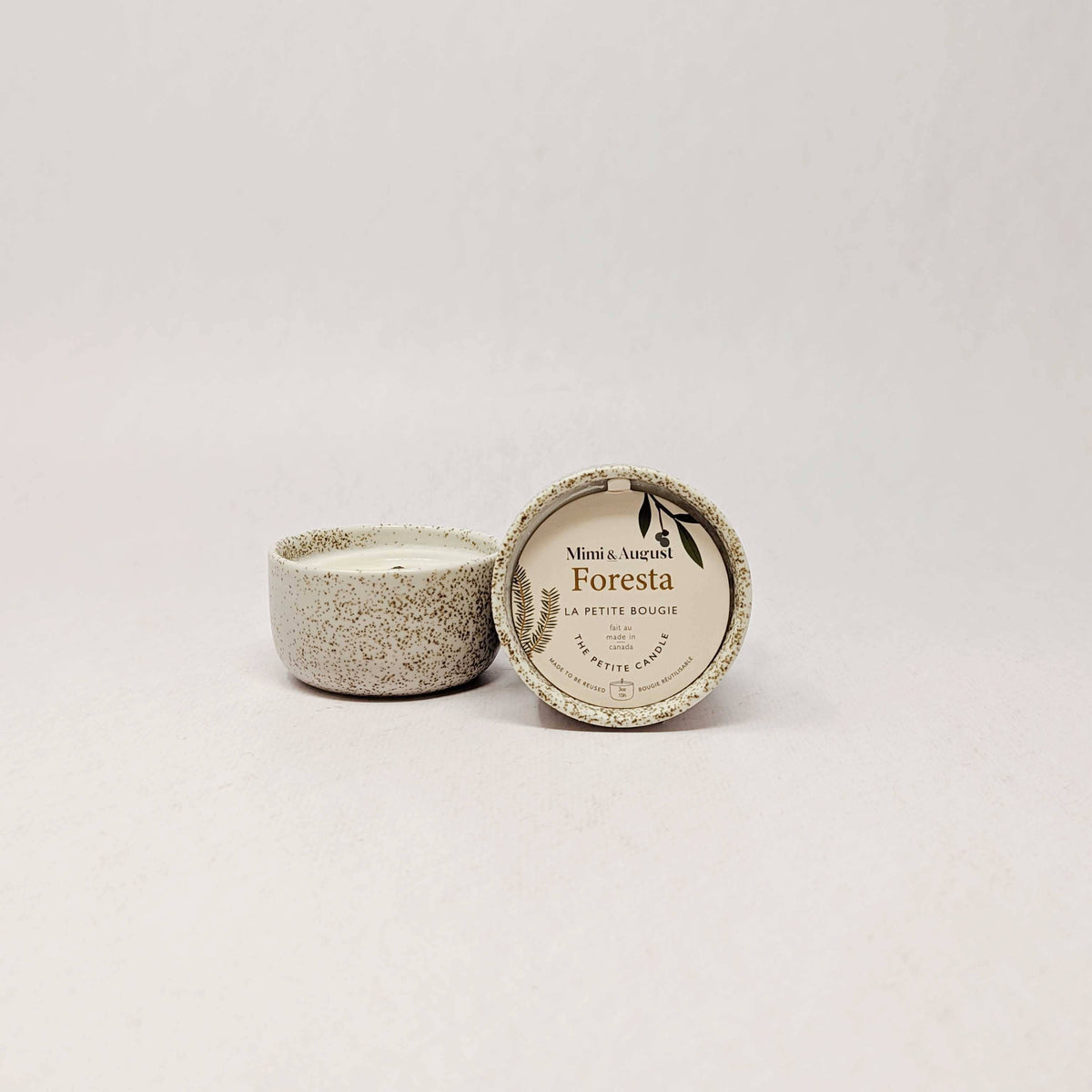 A small ceramic candle in a speckled beige pot with its lid leaning against it, showing a round label that reads 'Mint & Veuve Foresta' and 'La Petite Rouge'.