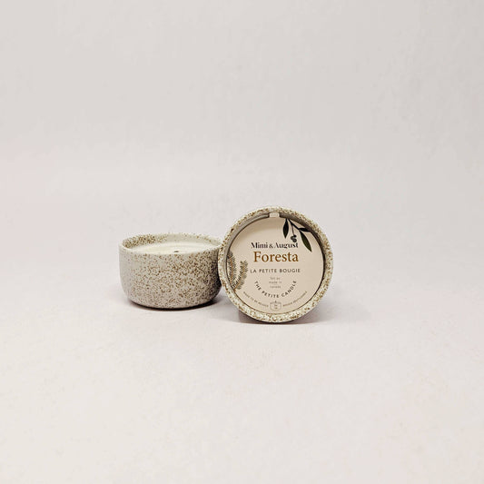 A small ceramic candle in a speckled beige pot with its lid leaning against it, showing a round label that reads 'Mint & Veuve Foresta' and 'La Petite Rouge'.