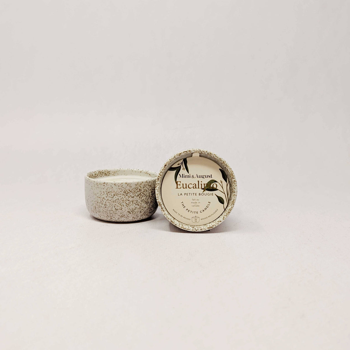 A small round ceramic candle tin with its lid off, showing a speckled beige and gray glaze. The lid is propped up next to the candle, which has a cream-colored label reading 'Eucalyptus' and 'La Petite Mâchique'.