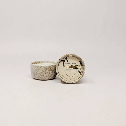 A small round ceramic candle tin with its lid off, showing a speckled beige and gray glaze. The lid is propped up next to the candle, which has a cream-colored label reading 'Eucalyptus' and 'La Petite Mâchique'.